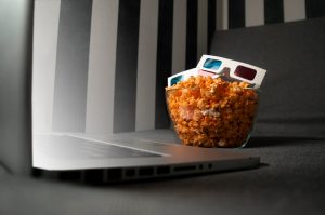 Computador sobre uma cama, com um pote de pipoca e óculos 3D para assistir filmes e séries relacionados a chatbots.